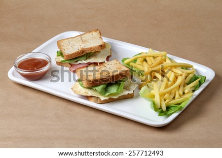 Club sandwich with ham and pork fried bacon, baked egg topped with cheese and lettuce next to a glass bowl of fresh hot red tomatoes ketchup and fine crispy baked french fries