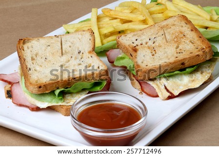 Club sandwich with ham and pork fried bacon, baked egg topped with cheese and lettuce next to a glass bowl of fresh hot red tomatoes ketchup and fine crispy baked french fries