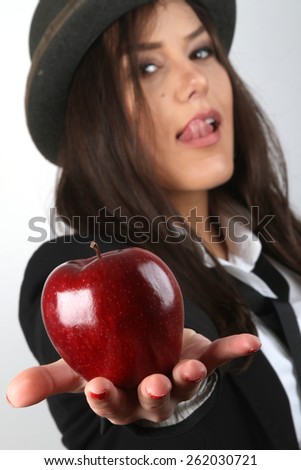 Girl and apple - Beautiful young brunette with red nail polish and a bowler hat on her head in a white shirt with a black tie in a black jacket holding a red organic fresh apple in hands provocatively