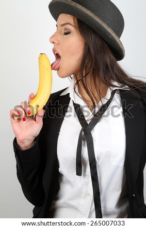 Beautiful girl and yellow banana - A beautiful brunette with gray old bowler hat on her head in a white shirt with a black jacket and black tie naughty play with fresh yellow raw banana