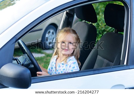 Happy little child, funny blonde toddler girl sitting of the car on driver seat holding steering wheel.