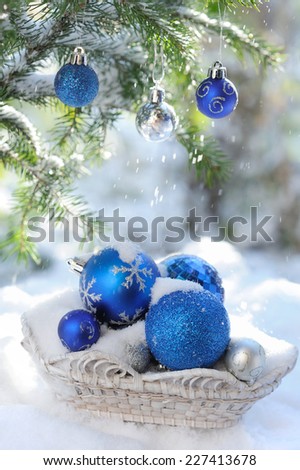 White basket with decorative xmas balls on the snow and blue balls on christmas tree outdoors in bright sunshine