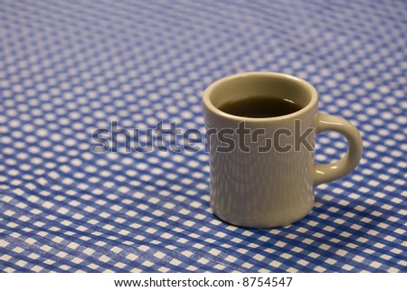 A plain mug of coffee on a plastic blue and white checkered tablecloth.