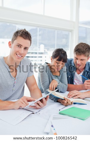 A smiling student using a tablet looks into the camera as his friends use a book to find the answer