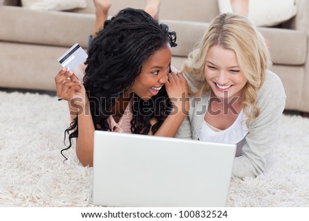 A woman lying on the floor is holding a bank card and smiling at her friend who has a laptop