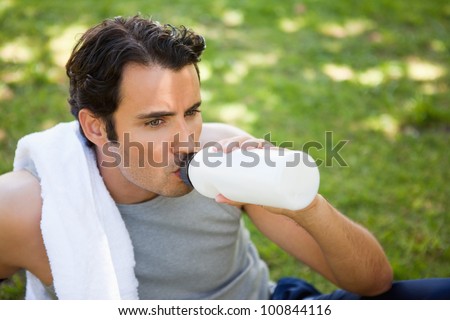Man with a white towel on his shoulder drinking from a sports bottle while sitting on the grass