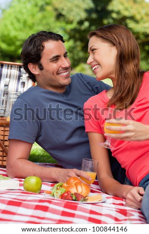 Two smiling friends looking into each others eyes while they hold glasses as they lie on a blanket with a picnic basket and food