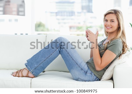 A woman sitting sideways on the couch, while looking forward and smiling with a mug in her hands.