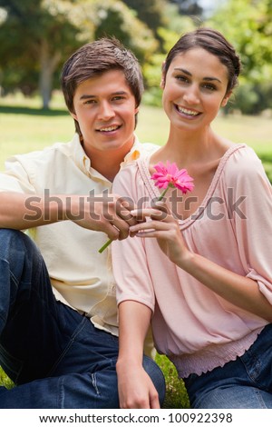 Man giving a flower to a woman as they both look straight in front of them while they sit on the grass