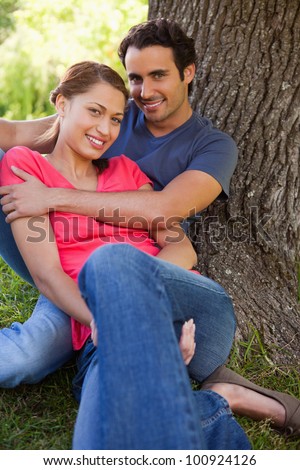 Man looking straight ahead as he holds his smiling friend while they sit together against the trunk of a tree