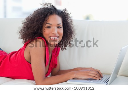 A smiling woman looking forward as she uses her laptop while lying forward on the couch.
