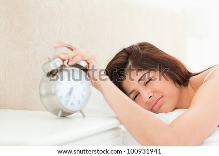 A woman is silencing the ringing bell of the alarm clock on the table beside the bed, with her hand. She is also only half awake.