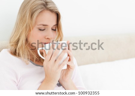 A woman with her eyes closed, in bed with a cup raised to her nose to take in the smell of the cups aroma.