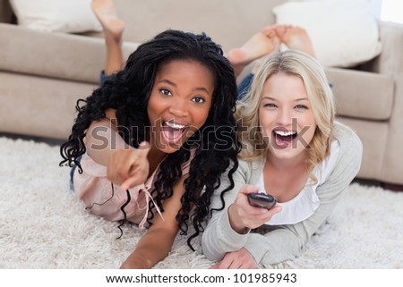 Two laughing women are lying on the ground and looking at the camera with a TV remote control