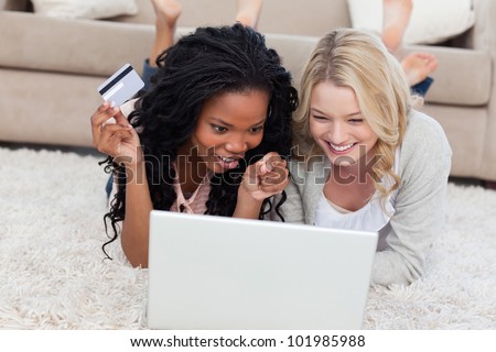 A woman has a bank card in her hand and is lying on the floor and looking at a laptop with her friend