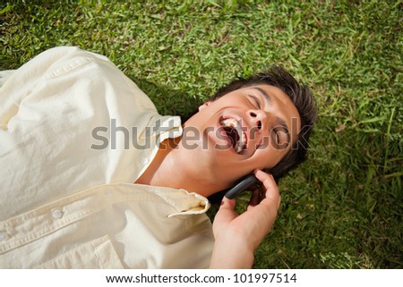 Man with his eyes closed laughing while making a call over the phone as he lies down on the grass