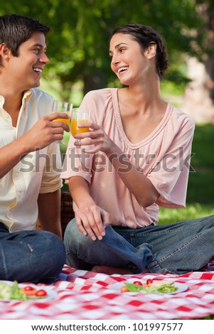 Woman laughing as she touches glasses of orange juice with her friend while they sit on a red and white picnic blanket