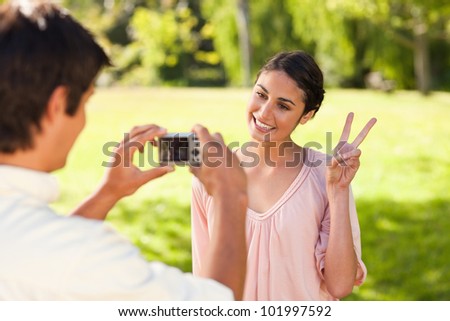 Using a camera, the man takes a photo of his friend smiling while giving the peace sign in a park