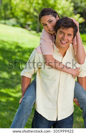 Woman smiling with her hand placed on her friends head as he is holding her on his back in a park