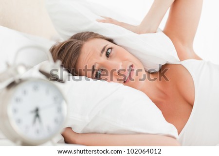 A woman uses a pillow to cover her ears as her alarm clock rings loudly.