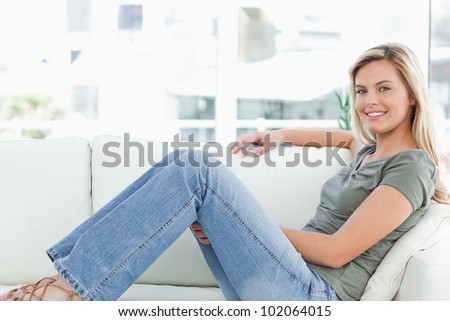 A woman with an arm across the top of the couch, sitting across the couch. smiling, legs raised and looking forward.