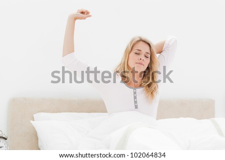 A woman sitting up in bed, with one arm stretched up, the other arm behind her head and her head looking forward, but with closed eyes.