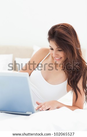 A close up shot of a woman lying on the bed as she smiles while she uses her laptop.