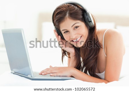 A woman looking forward and smiling as she uses a laptop and listens to headphones as she relaxes on the bed.