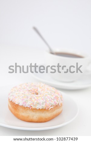 Doughnut with multi coloured icing sugar and a cup of coffee with a spoon on white plates against a white background
