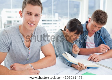A male student looking into the camera and smile as the other students sit and look at their books