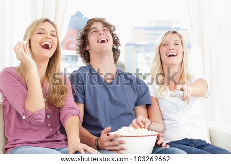 Three friends eating popcorn while laughing at the show while they sit on the couch together