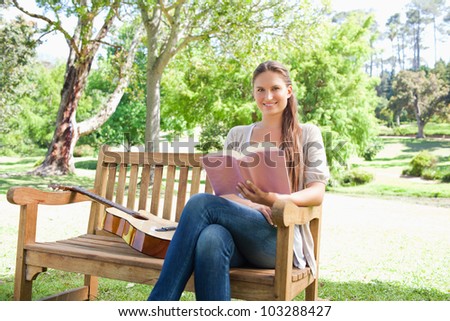 Smiling young woman with a book and a guitar sitting on a bench