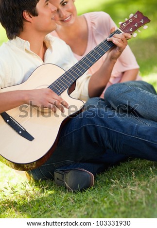 Man playing a song on the guitar while his smiling friend watches and listens to him as they sit on the grass