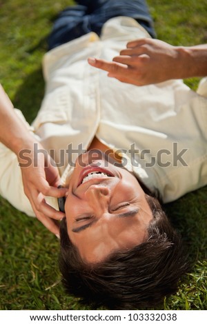 Man with his hand raised laughing while making a call over the phone as he lies down on the grass