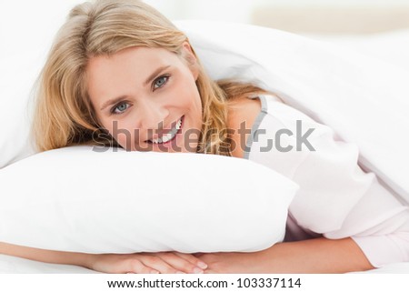 A woman lying in bed, with her head raised from the pillow slightly, smiling as her hands are under the pillow.