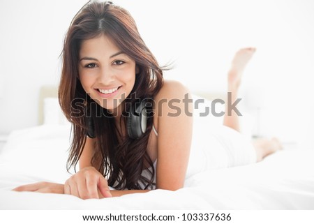 A woman with headphones on, lying on a bed while raising one leg upwards, and smiling.