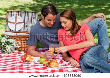 Two friends touching glasses against each other in celebration while lying on a blanket with picnic food