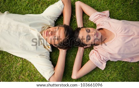 Man and a woman with their eyes closed lying head to head with both of their arms resting behind their neck on the grass