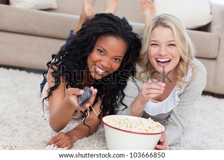 Two women lying on the ground with popcorn are smiling at the camera nd holding a TV remote