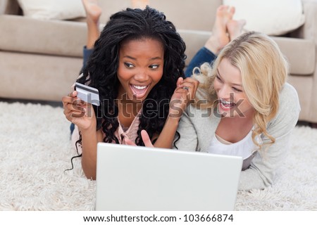 Two women are laughing and lying on the floor with a laptop and bank card