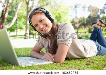Smiling young woman with a headset and a laptop lying on the lawn