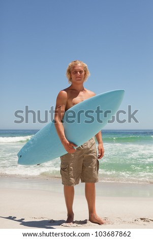 Blonde man holding his blue surfboard while standing on the beach