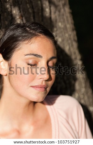 Woman sitting against the trunk of a tree with her eyes closed as the sun shines on her face