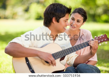 Man playing a song on the guitar while his smiling friend watches him as they are both sitting on the grass