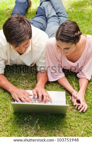 Two friends smiling while watching something on a laptop as they lie down together in the grass