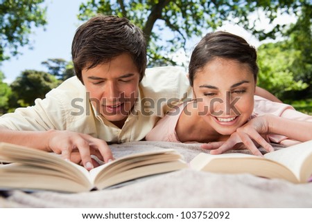 Man and a woman smiling while reading books together as they are lying prone on a blanket in the grass