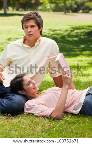 Woman smiling while she is lying against her friend's leg as she is reading a book
