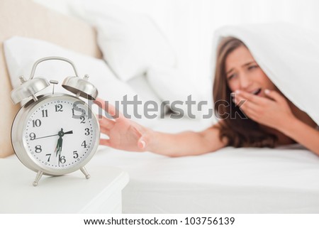 A woman is reaching out to silence her alarm clock while underneath her blanket in bed.