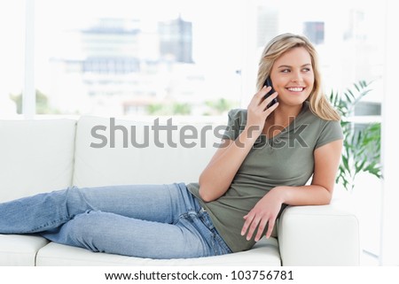 A woman smiling and looking to the side, while she makes a call and lies across the couch.