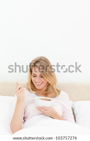 A vertical shot of a woman in bed with a bowl of cereal, as she smiles and looks at the spoon of cereal.